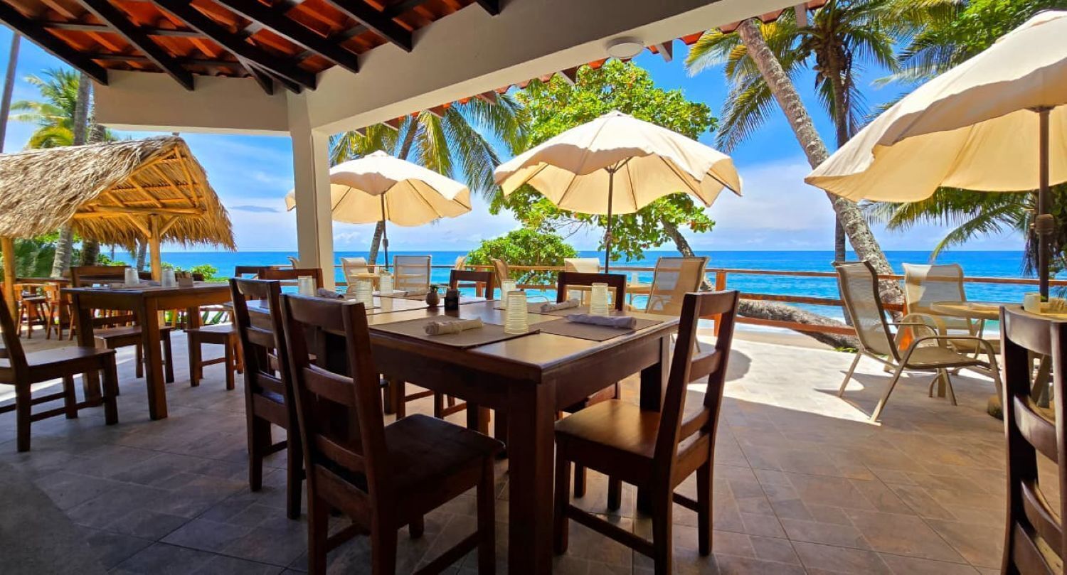 Un espace extérieur aménagé pour les repas, avec des tables et des chaises en bois, des parasols de couleur beige et une vue sur l’océan.