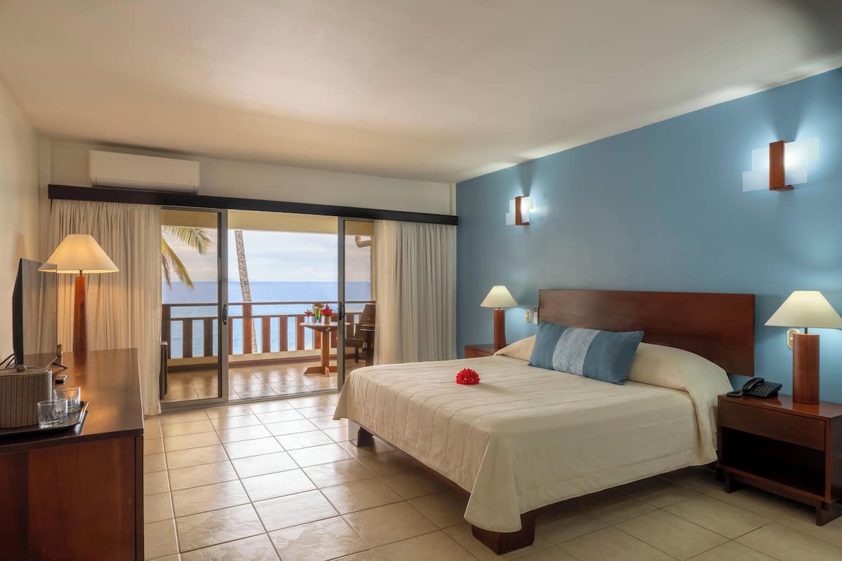 Une chambre d’hôtel comprenant un lit, des tables de chevet, un bureau avec une lampe, une chaise et une vue sur l’océan.