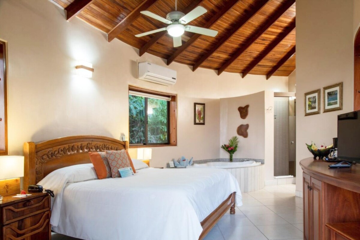 A bedroom with a wooden ceiling, a bed, a nightstand, a desk, a vanity, a bathtub, and a shower.
