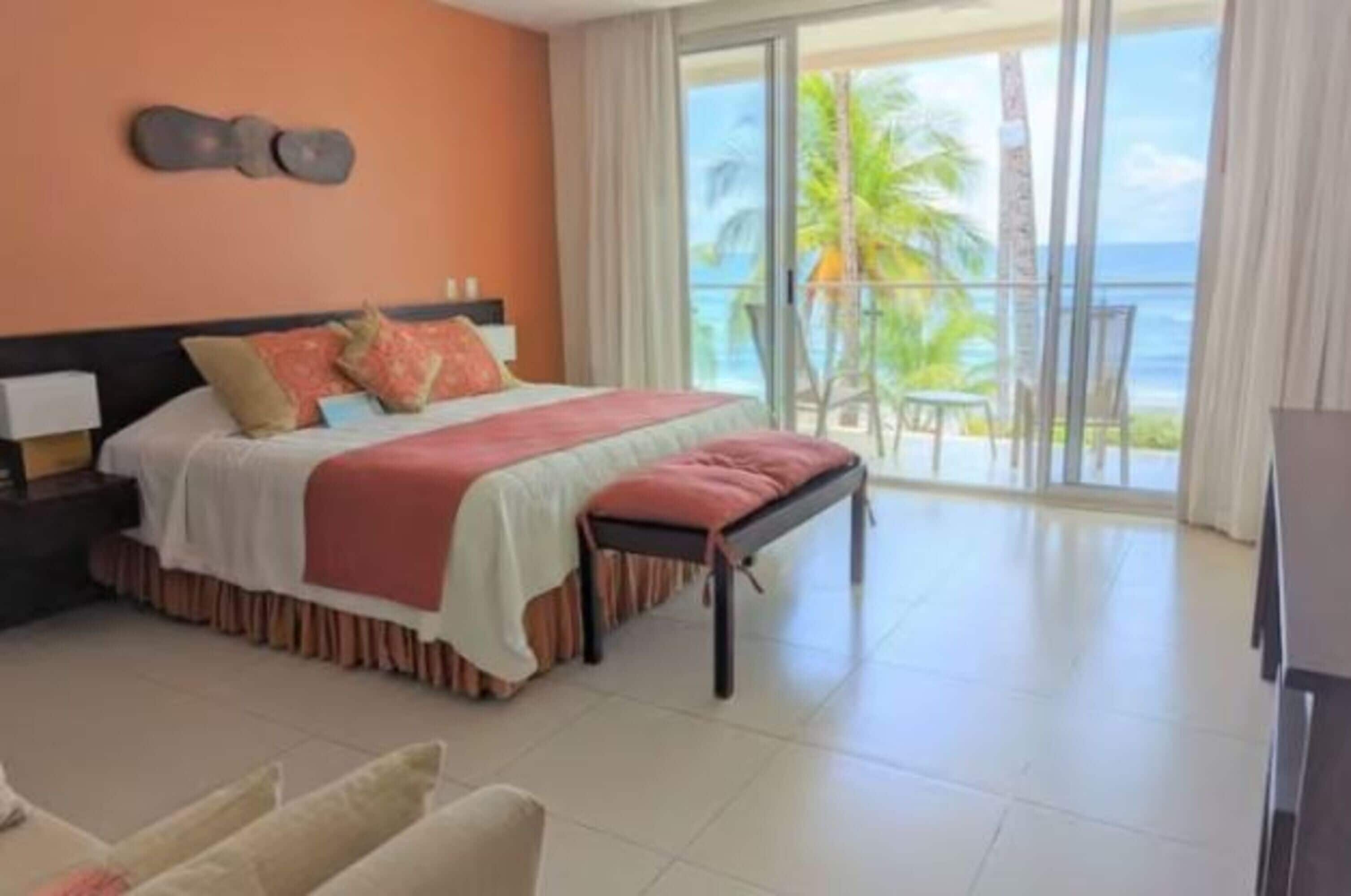 Quarto de hotel com cama, mesa de cabeceira, poltrona e vista do oceano através de grandes janelas.