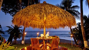 A wooden deck with a thatched umbrella table set for two, overlooking a beach.
