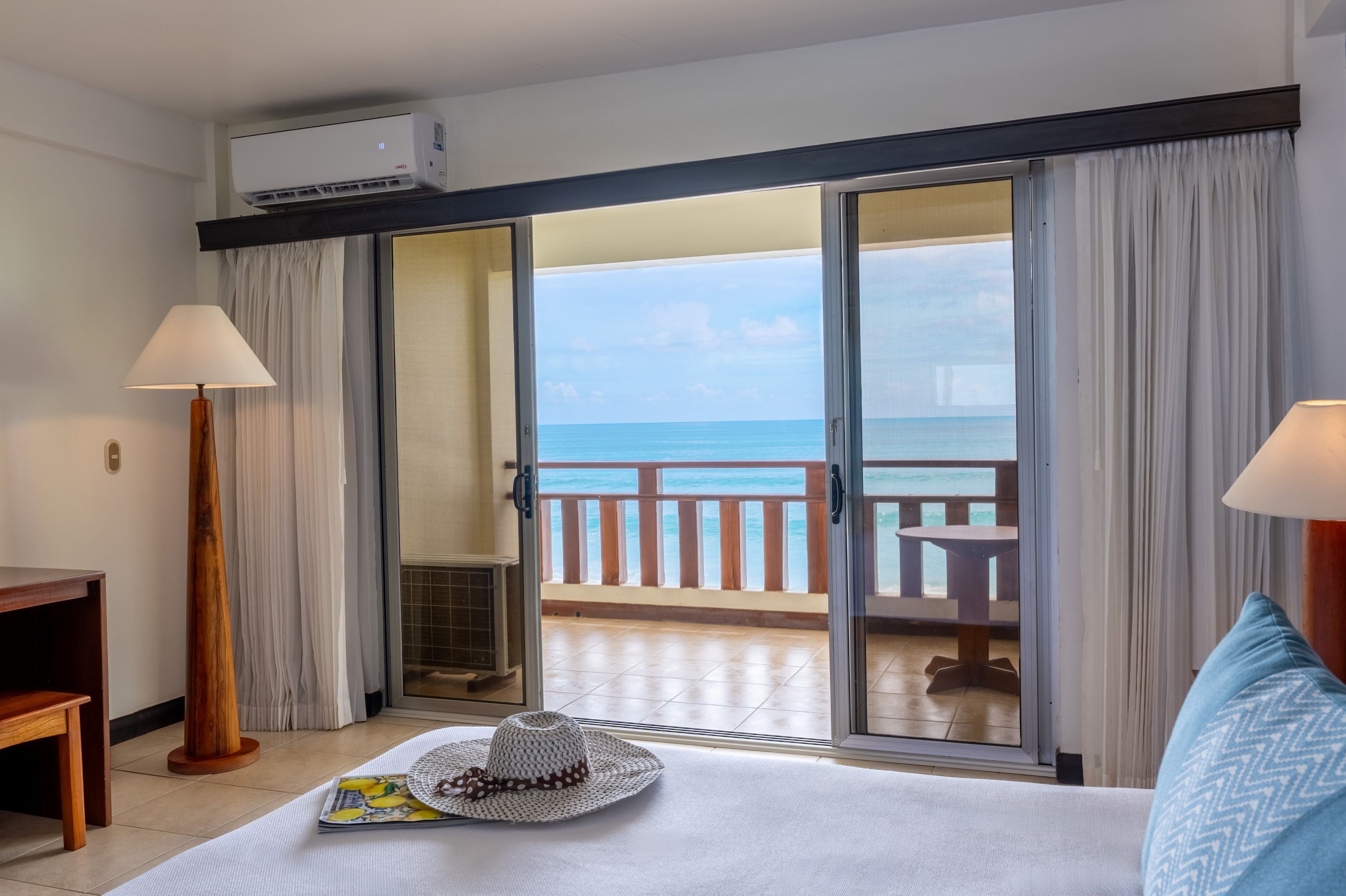 Una camera d'albergo con un letto, una scrivania, una lampada, un cappello e vista sull'oceano attraverso una porta scorrevole.
