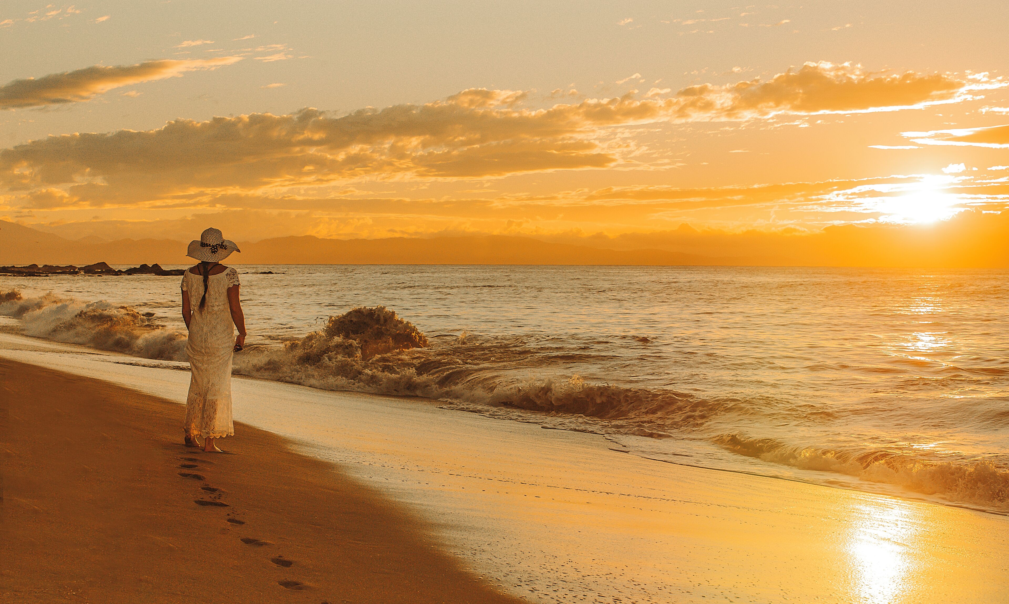 Una playa con una persona que camina por la orilla, vestida de blanco y con sombrero.