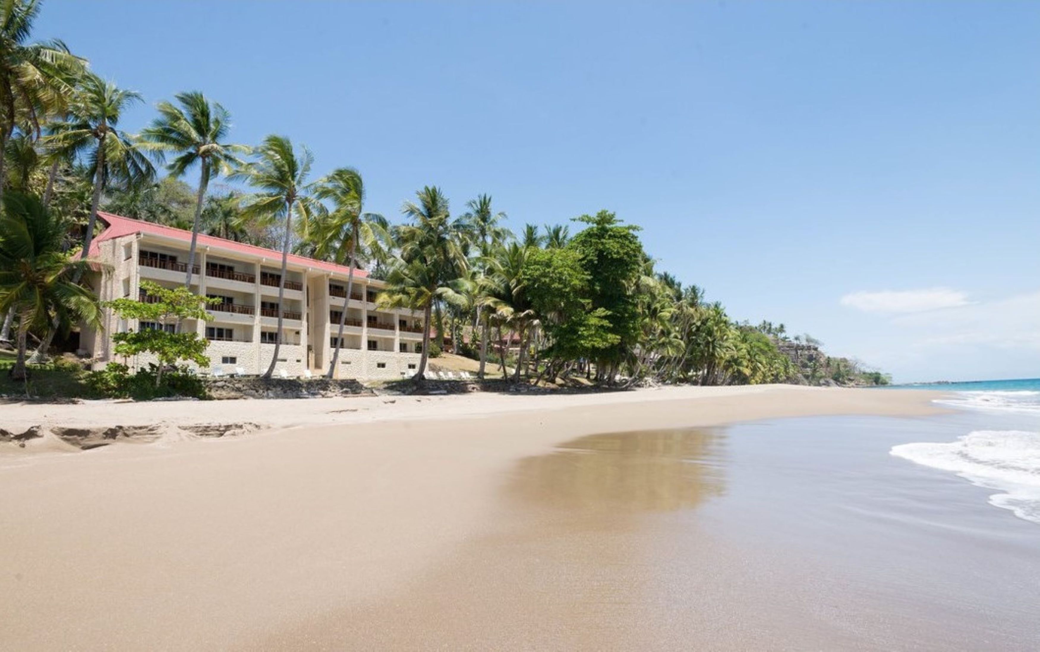 A beachfront hotel with multiple floors, palm trees, and a sandy beach.