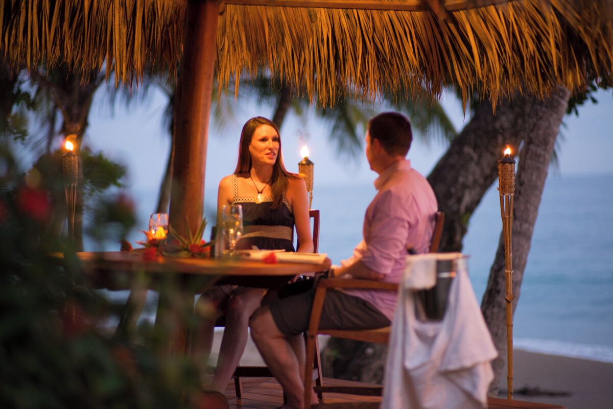 A couple dining at a table under a thatched roof, with a candlelit ambiance and a view of the ocean.