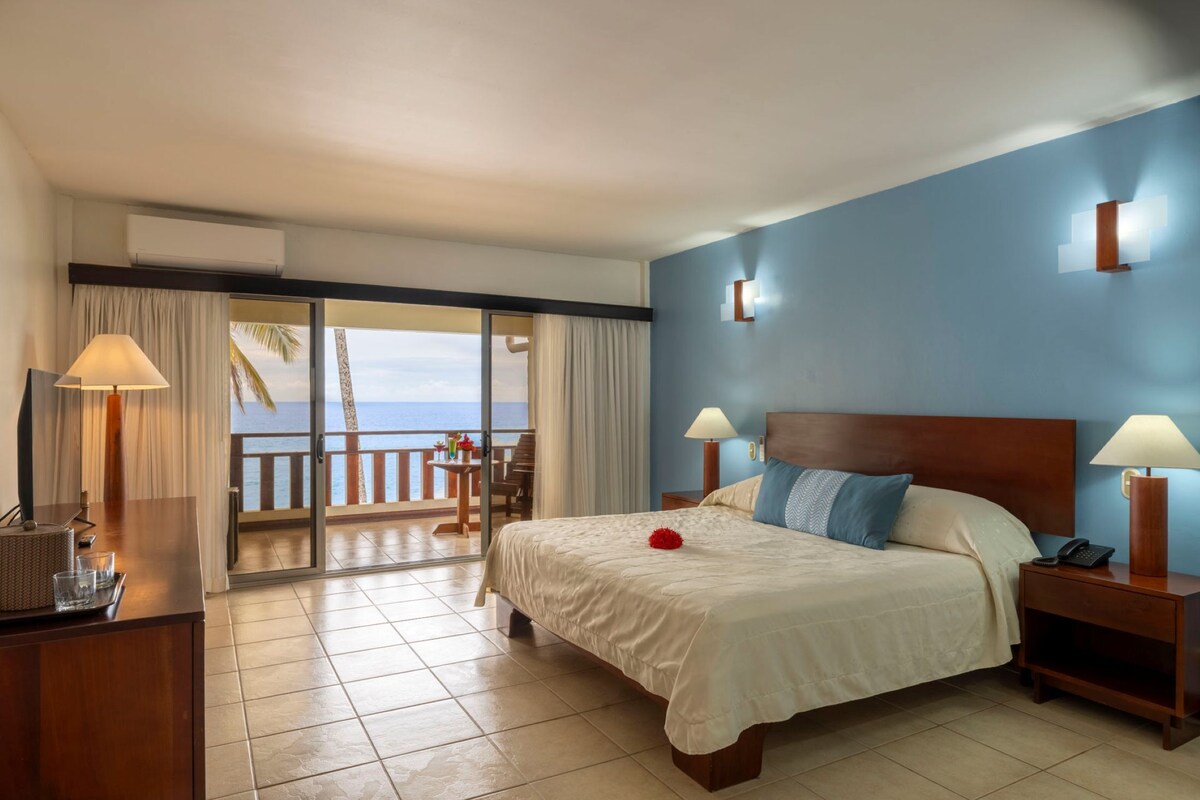 A hotel room with a bed, bedside tables, a lamp, a view of the ocean, and a balcony with a table and chairs.