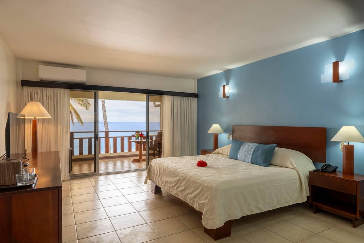 Quarto de hotel com cama, mesas de cabeceira, abajur, vista para o oceano e varanda com mesa e cadeiras.