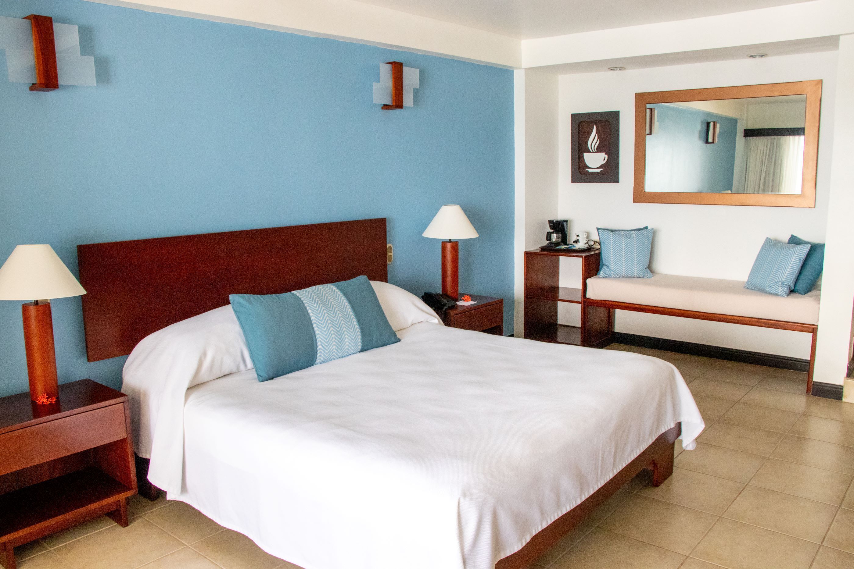 Quarto de hotel com cama, luminárias de cabeceira, um banquinho, um espelho e uma parede com arte de uma xícara de café.
