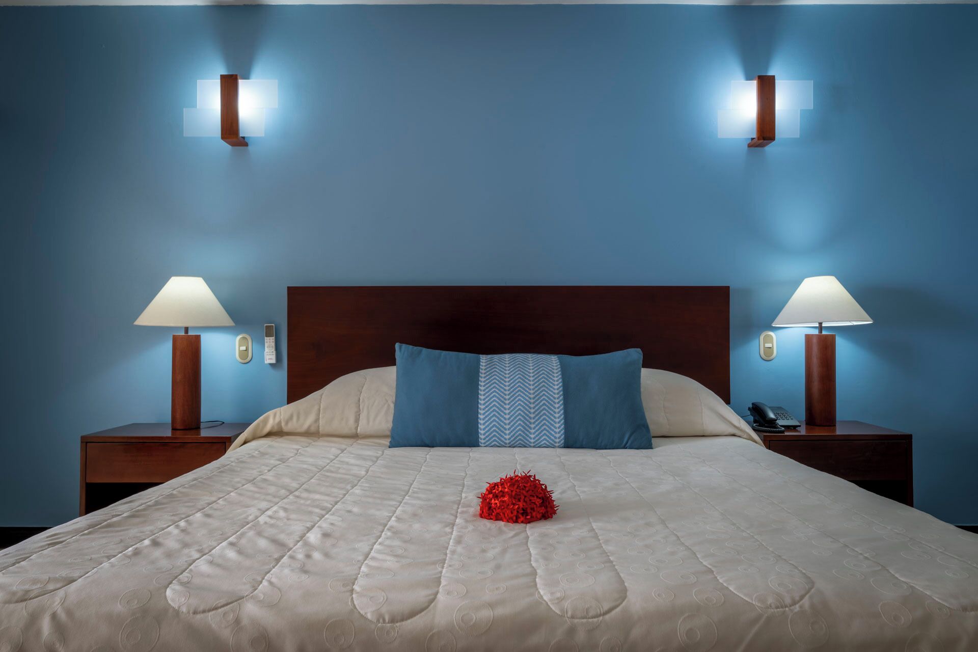 A neatly made bed with a blue pillow, two bedside lamps, and a bedside phone on a wooden nightstand.