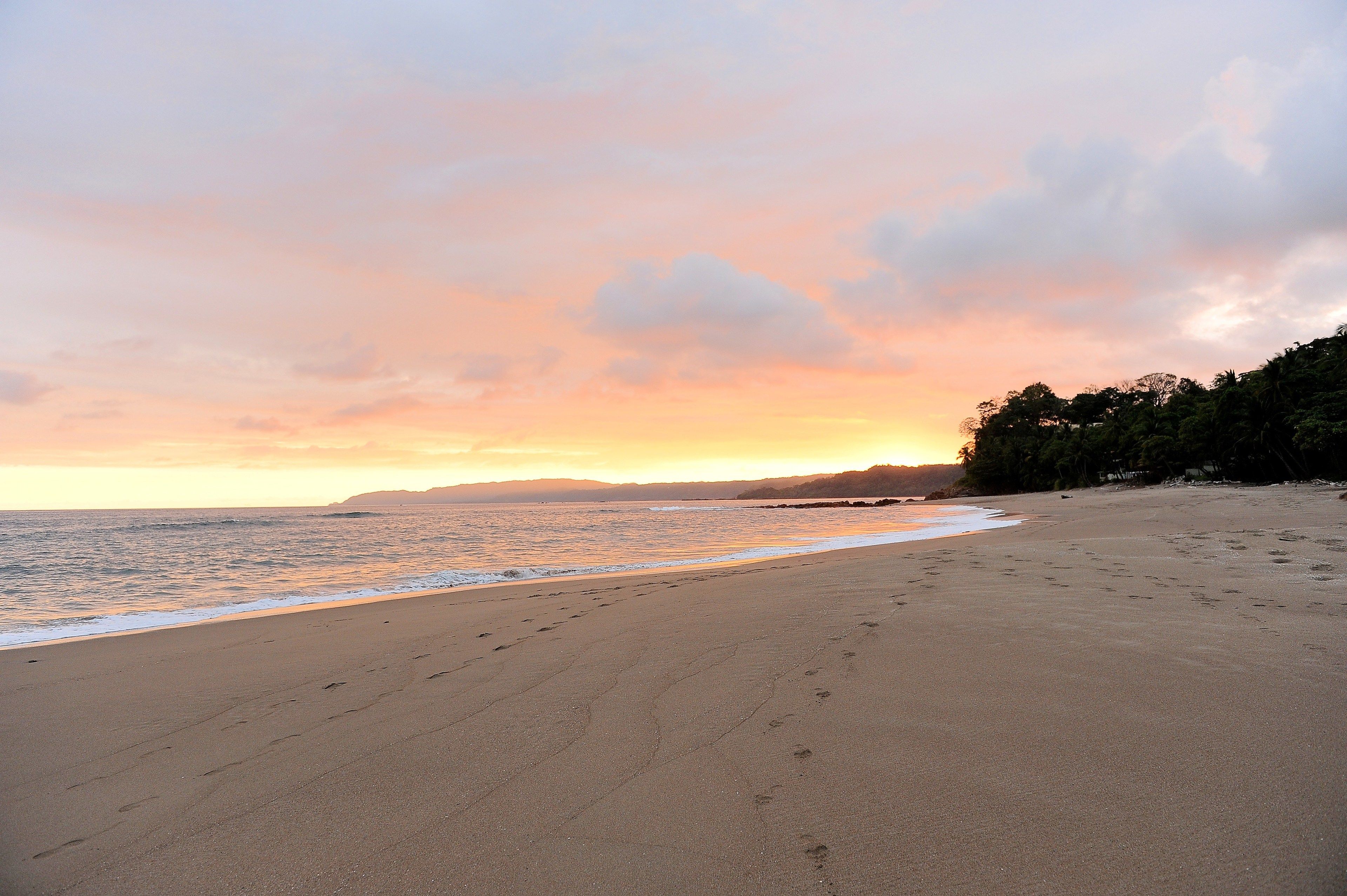 A beach with footprints leading to the water, a clear sky, and a distant mountain range.