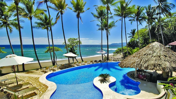 A pool with a unique design, surrounded by lounge chairs and umbrellas, with a beach and palm trees in the background.