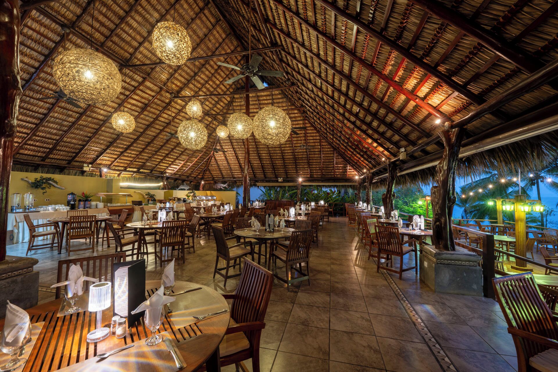 Un espaiós restaurant amb un alt teulat de palla, diverses taules de menjador i lluminàries esfèriques penjants.