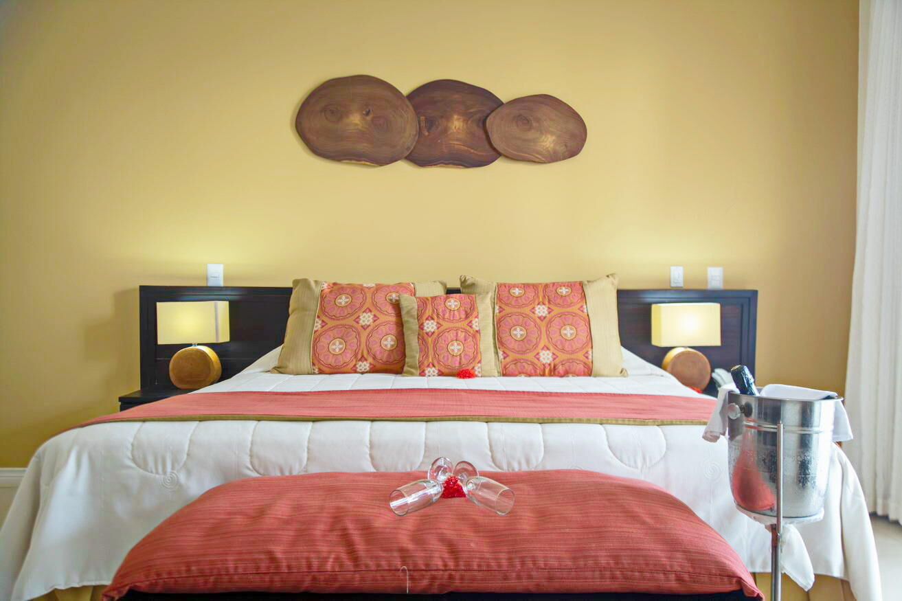A hotel room with a bed, pillows, bedside lamps, a nightstand, and a bedside table with a lamp and a bottle.