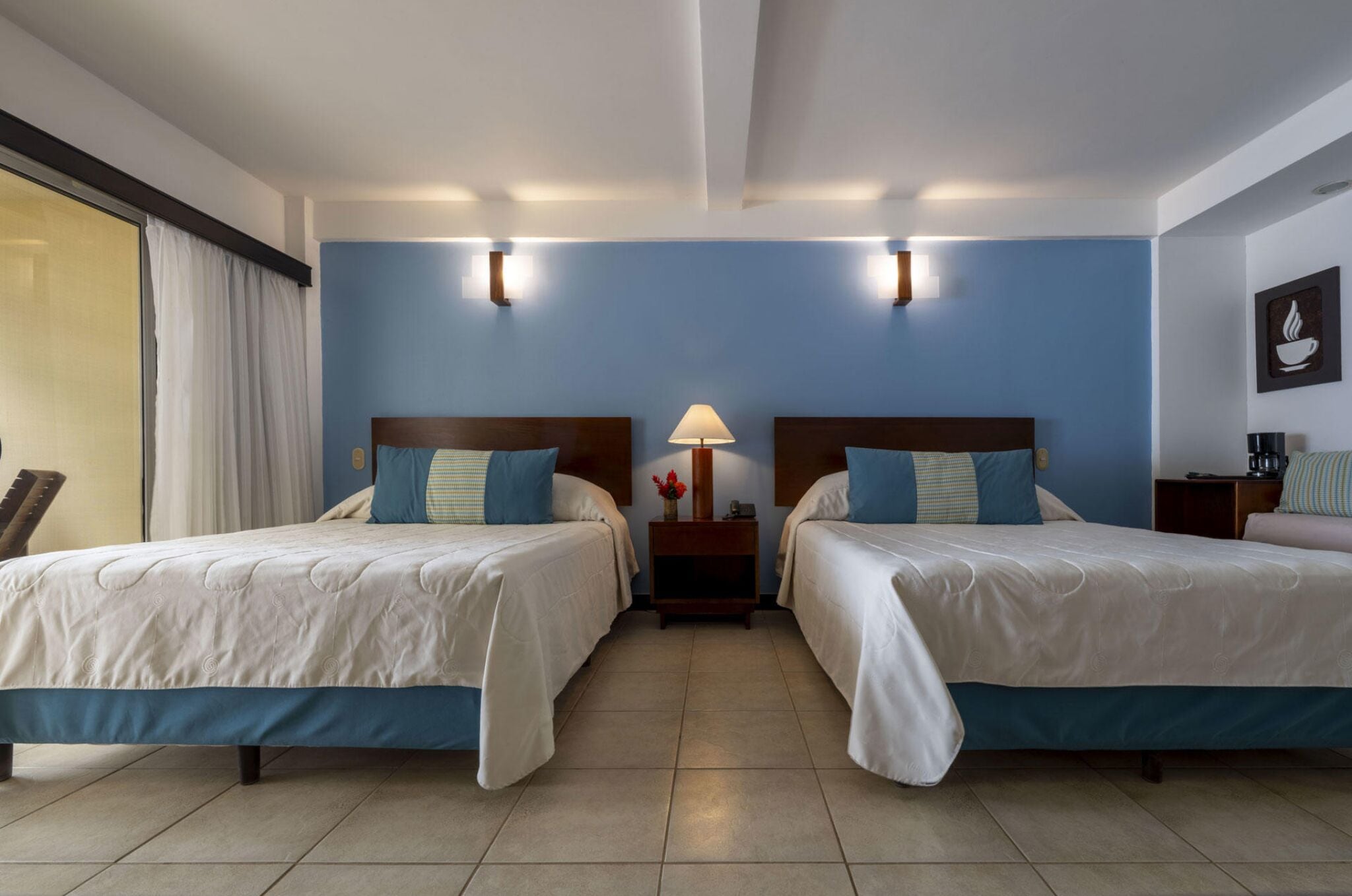 Quarto de hotel com duas camas, uma mesa de cabeceira com abajur e luminária na parede.