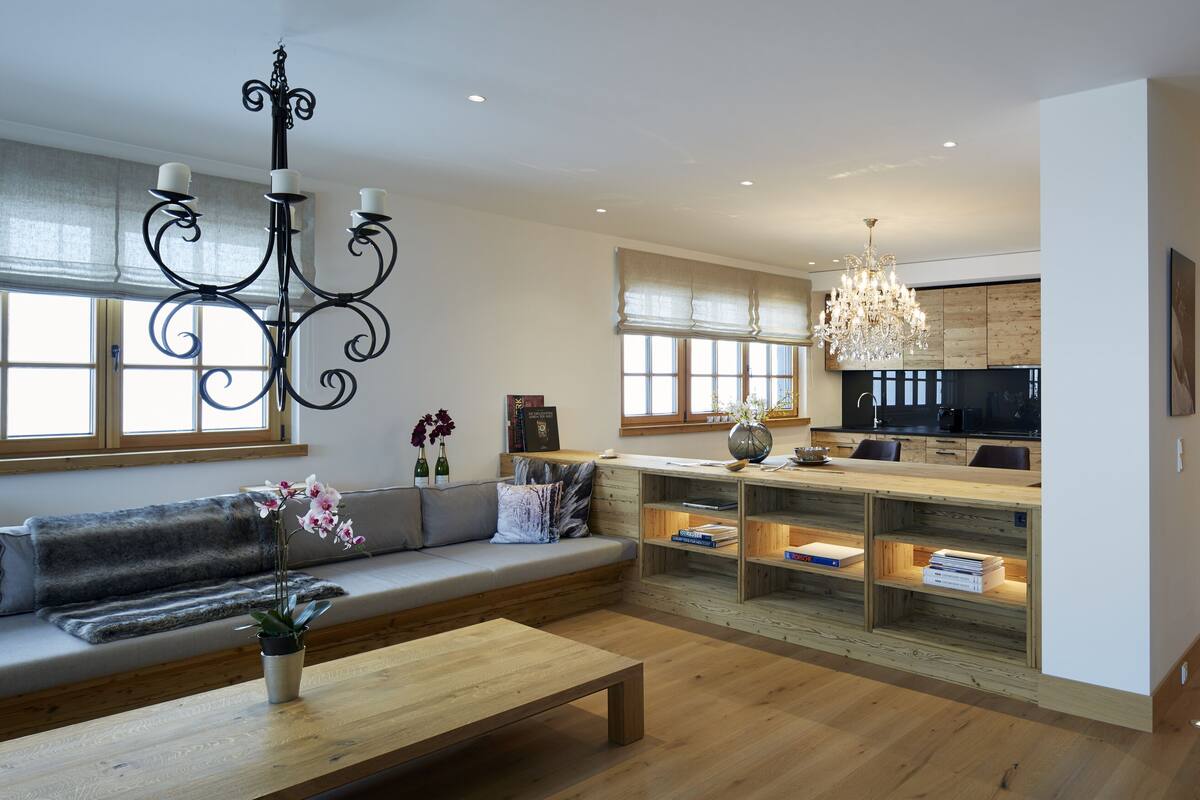 Ein modernes Wohnzimmer mit einer Couch, einem Couchtisch und dekorativen Elementen. Im Hintergrund ist eine Küche mit Holzschränken und einer Chandelier zu sehen.