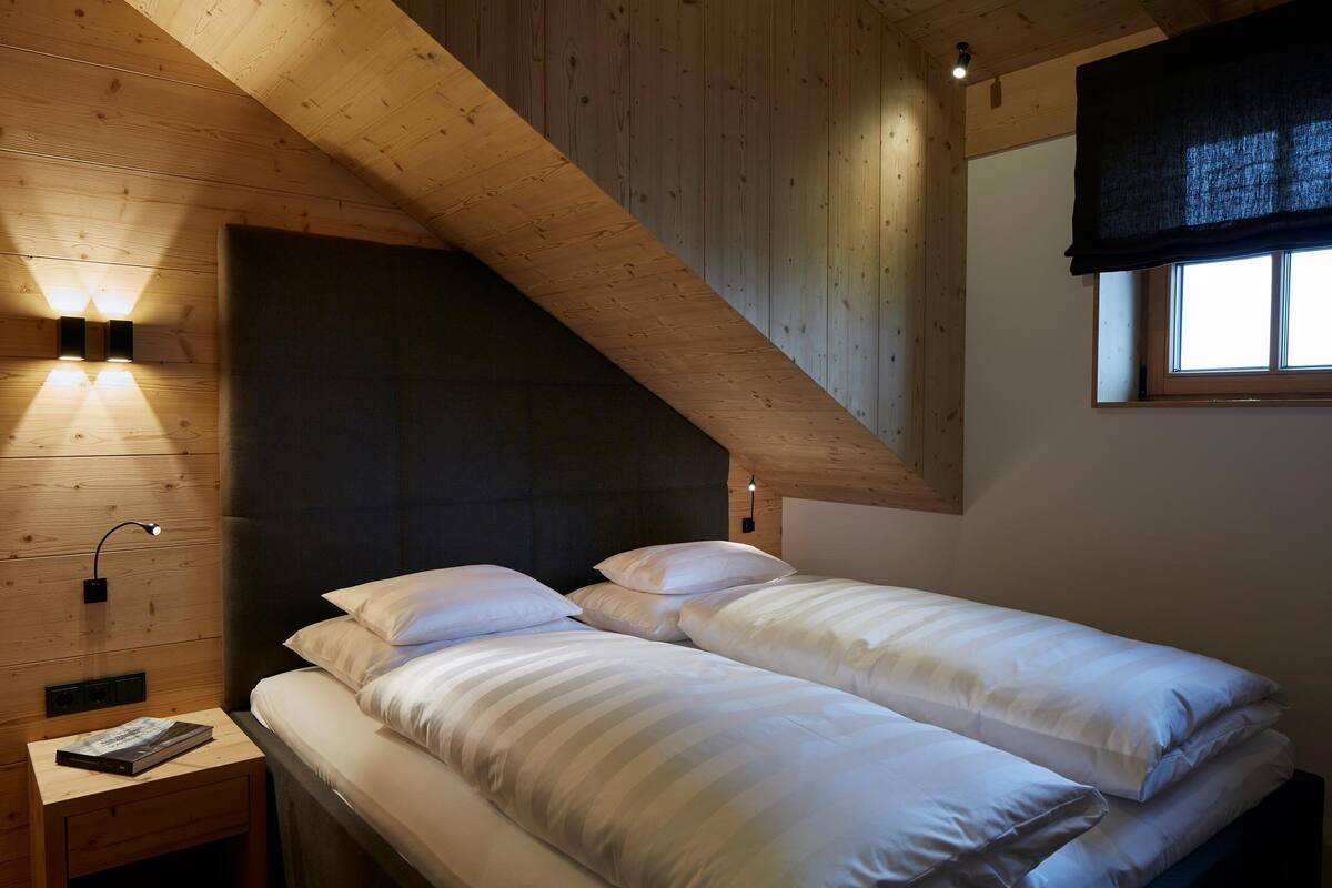 Ein Schlafzimmer mit Holzboden, einem Bett mit weißer Bettwäsche, einem Nachttisch mit Lampe und einem Fenster mit Verdunkelungsvorhang.