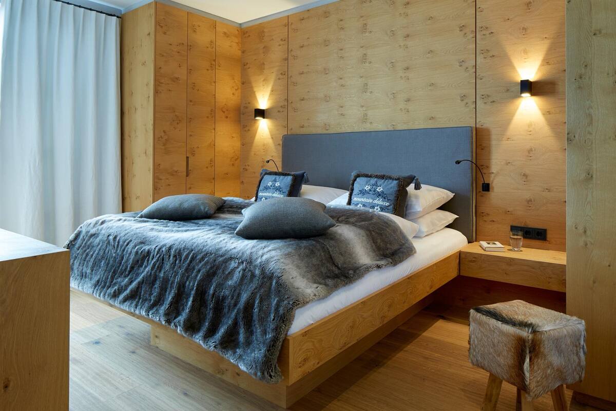 Ein modernes Schlafzimmer mit einem Holzkopfteil, einem großen Bett mit grauer Bettwäsche, zwei Wandleuchten und einem kleinen Holzschemel.