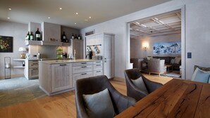A modern kitchen with wooden cabinets, a dining area with a wooden table and leather chairs, and a living space with a sofa and a painting of mountains. - Arlberg Hospiz Chalet Suiten (Sankt Anton am Arlberg)