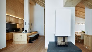 A modern kitchen with wooden cabinets, a central island with bar stools, and a built-in fireplace. - Arlberg Hospiz Chalet Suiten (Sankt Anton am Arlberg)