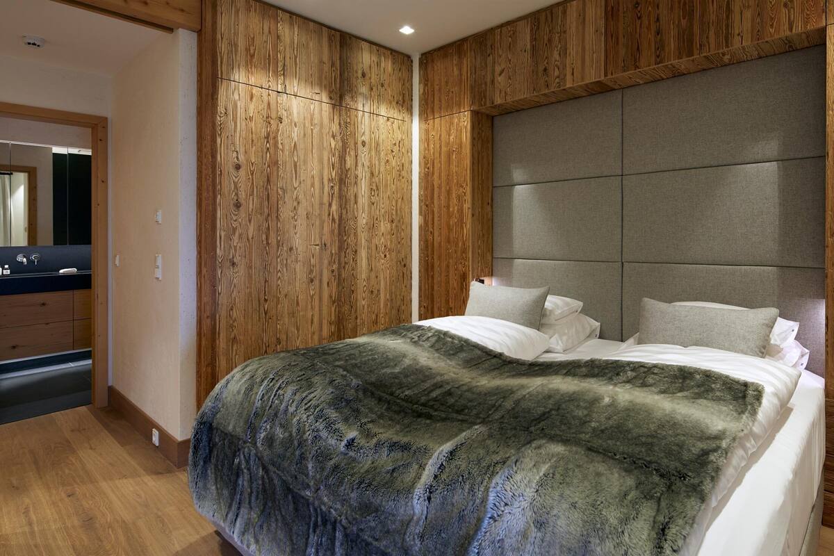 Ein modernes Schlafzimmer mit einer Holzwand, einem großen Bett mit grauem Kopfteil und einem Badezimmer mit einem Holzwaschtisch.