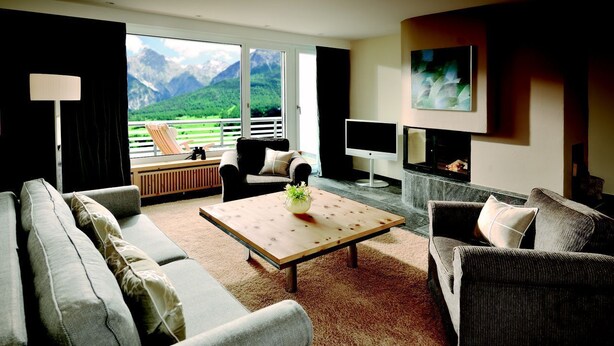 A living room with a sofa, armchairs, a coffee table, a TV, and a fireplace. There is a balcony with a view of mountains. - Hotel Paradies (Ftan)