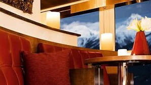 A red leather sofa with a single red cushion, a small round table with a vase, and a view of clouds through a window. - Hotel Paradies (Ftan)