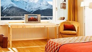 A hotel room with a bed, a chair, a window with a mountain view, a radiator, and a lamp. - Hotel Paradies (Ftan)