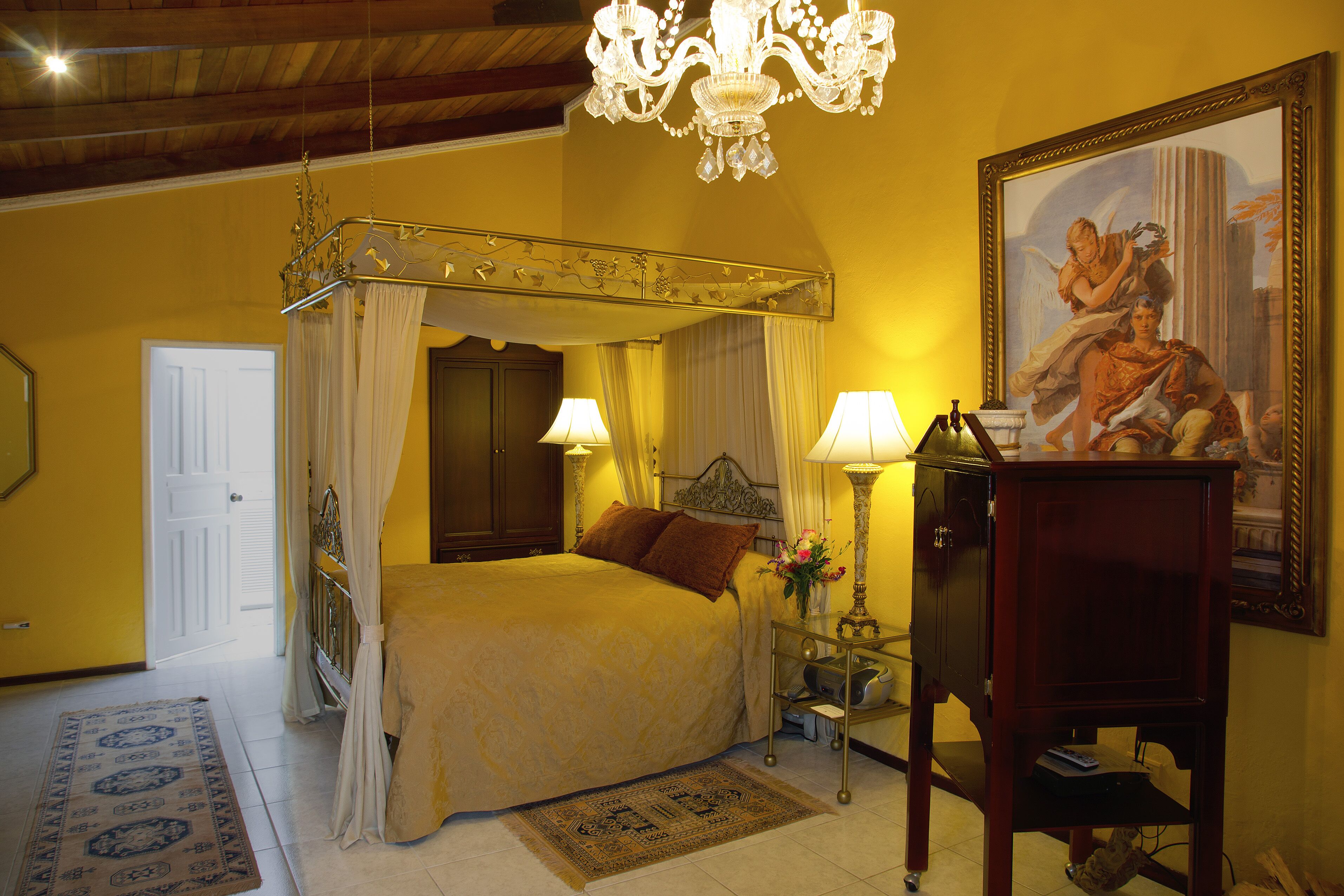 A bedroom with a four-poster bed, a chandelier, a painting, and a nightstand with a lamp.