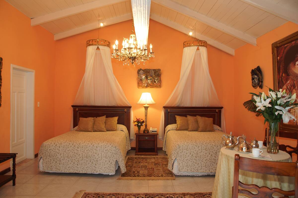 Quarto de hotel com duas camas, um candelabro, uma mesa com chá e um quadro na parede.