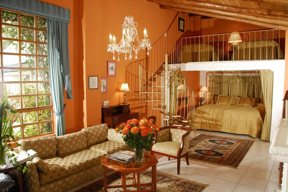 Um quarto com sofá, mesa com um vaso de flores, lustre, escada e cama com colcha estampada.