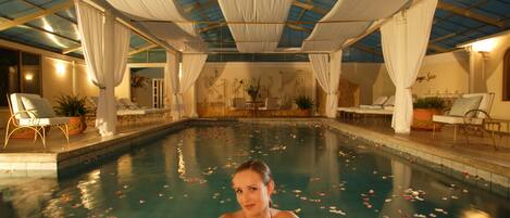 A pool area with a glass ceiling, white curtains, and a woman sitting by the pool.