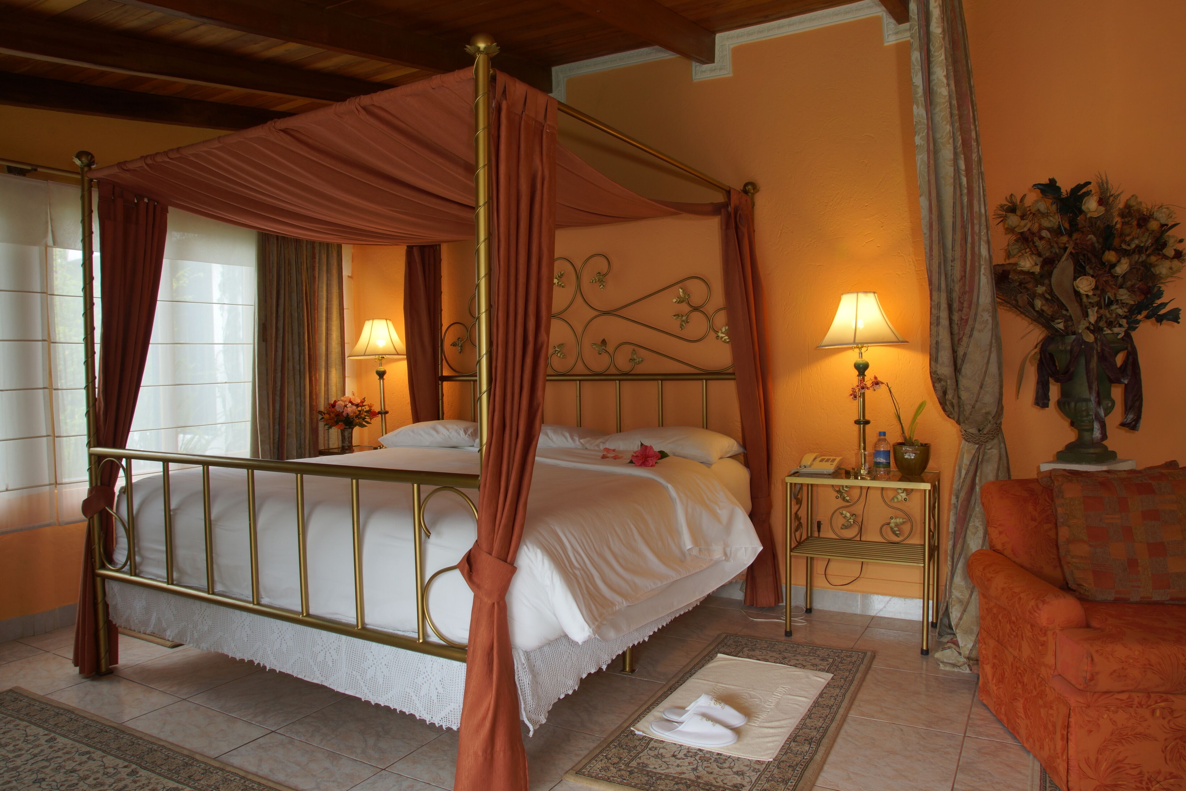 A four-poster bed with a canopy, two bedside lamps, a small table with a vase of flowers, and an orange armchair.