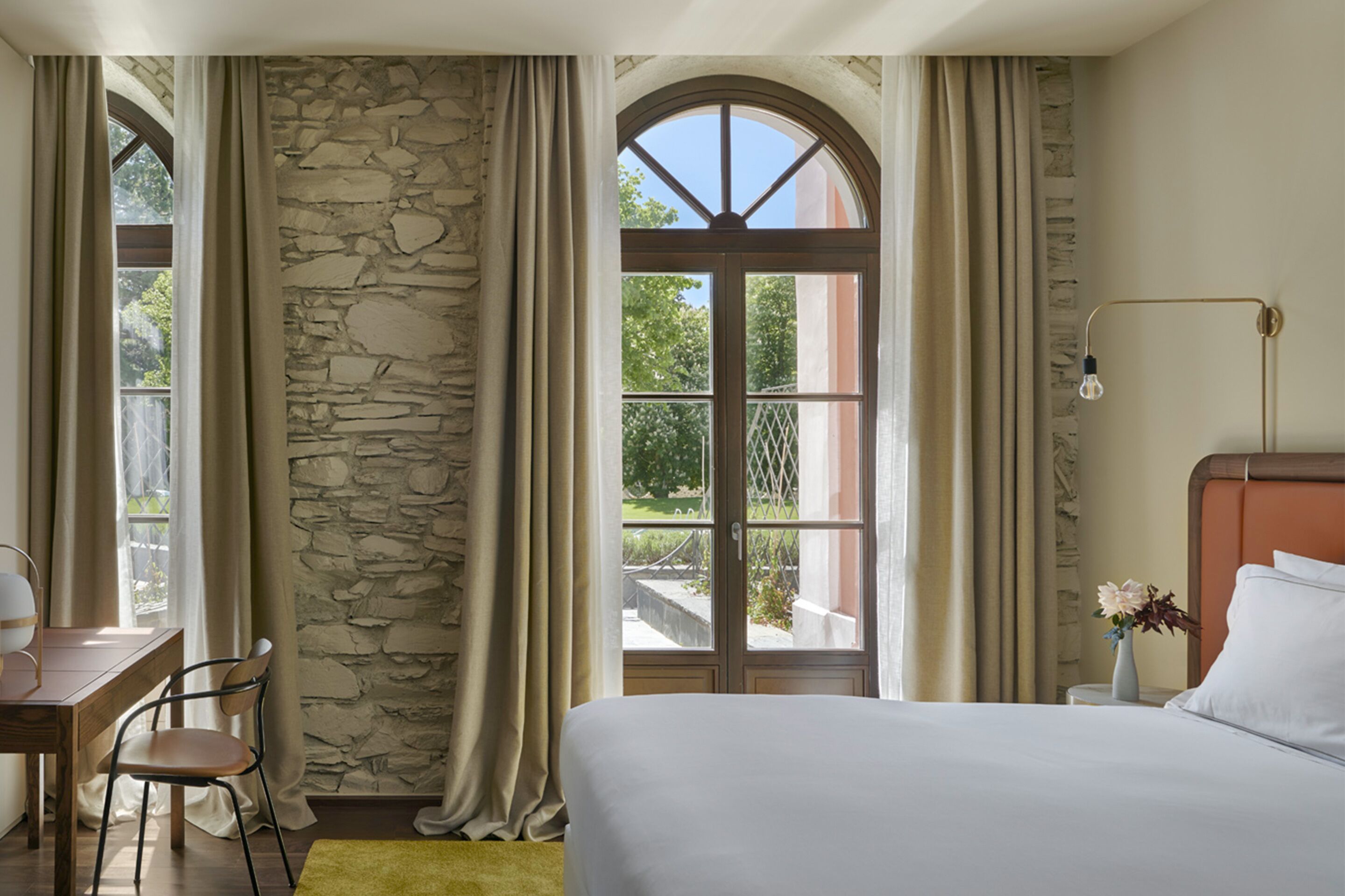 Un dormitorio con pared de piedra, un amplio ventanal de arco, cama, mesita de noche, lámpara y una silla.