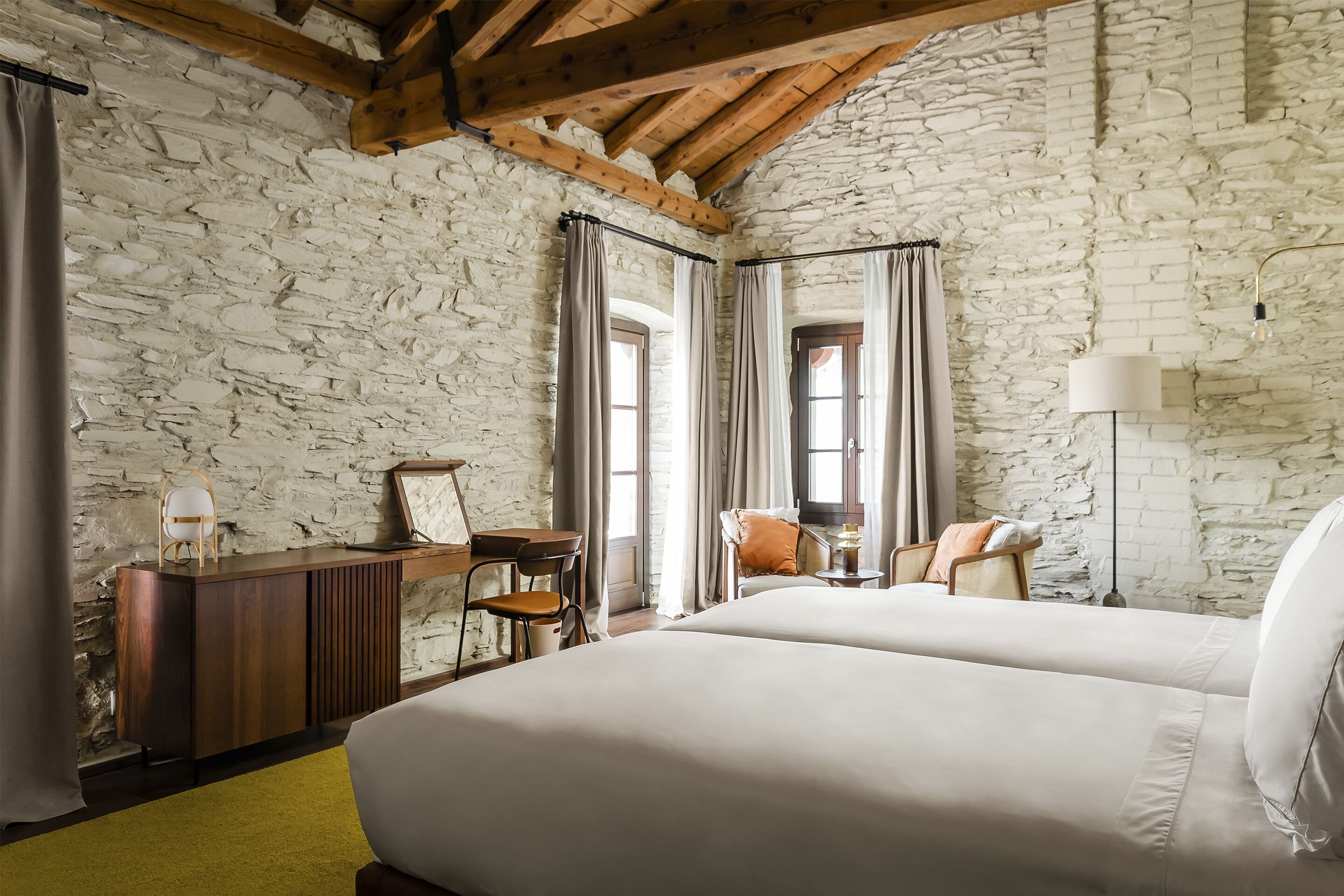 A room with a stone wall, wooden beams, a bed, a desk, and a chair.