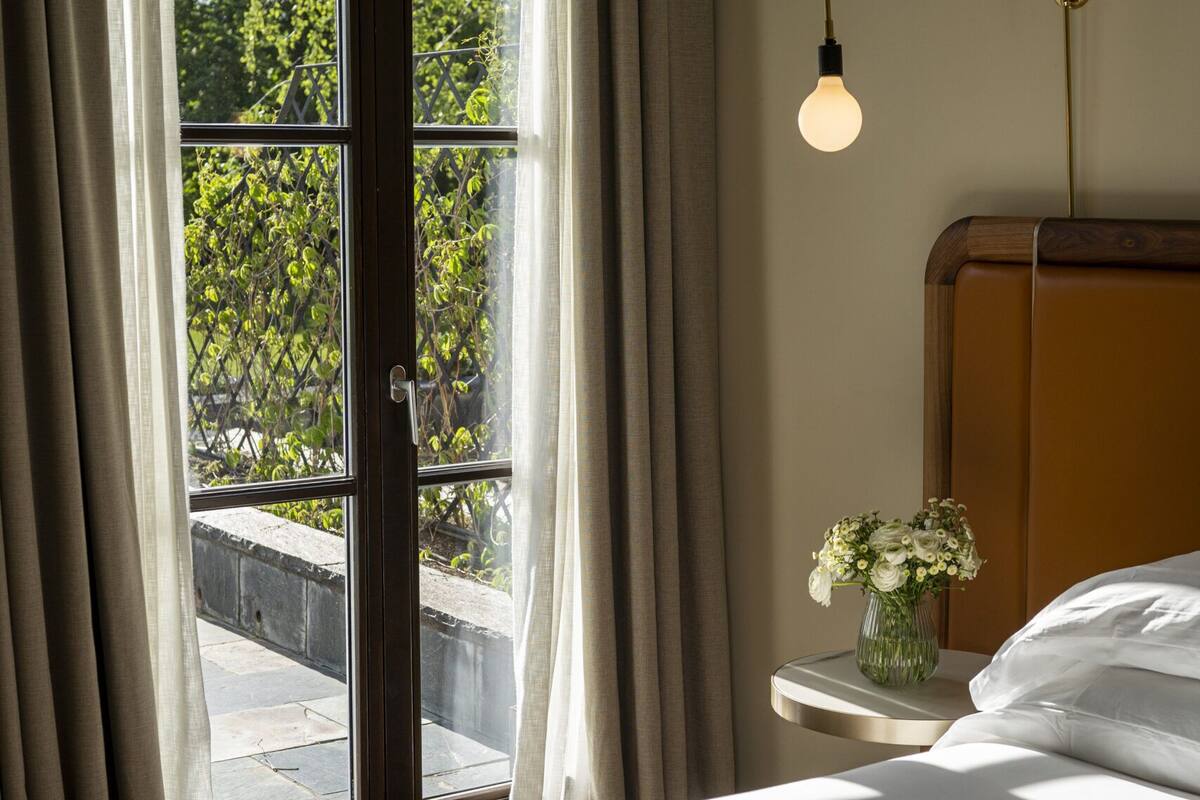 A bedroom with a bed, a nightstand with a vase of flowers, a window with curtains, and a wall-mounted lamp.