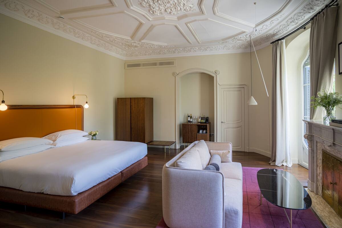 A hotel room with a large bed, a sofa, a small table, and a fireplace.