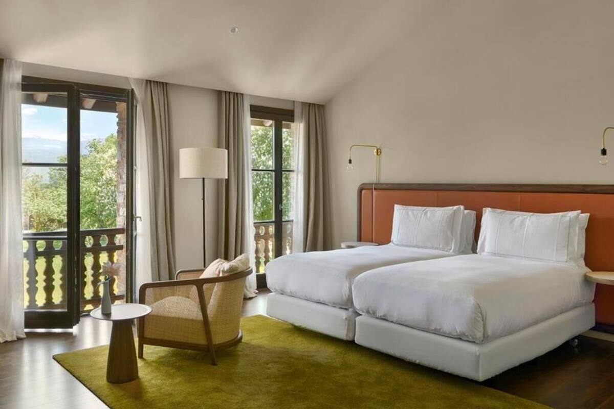 A hotel room with two beds, a chair, a small table, and a view of greenery through large windows.