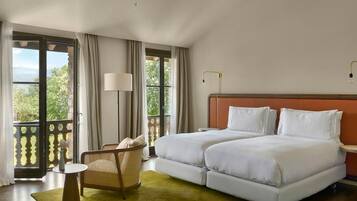 A hotel room with two beds, a chair, a small table, and a view of greenery through large windows.