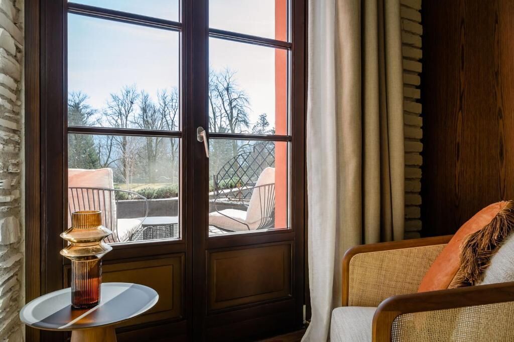 A room with a large window, a small table with a decorative vase, and a wicker chair.