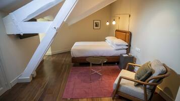 Een kleine, knusse slaapkamer met een schuine kap, een enkel bed, een stoel, een klein tafeltje en een wandlamp.