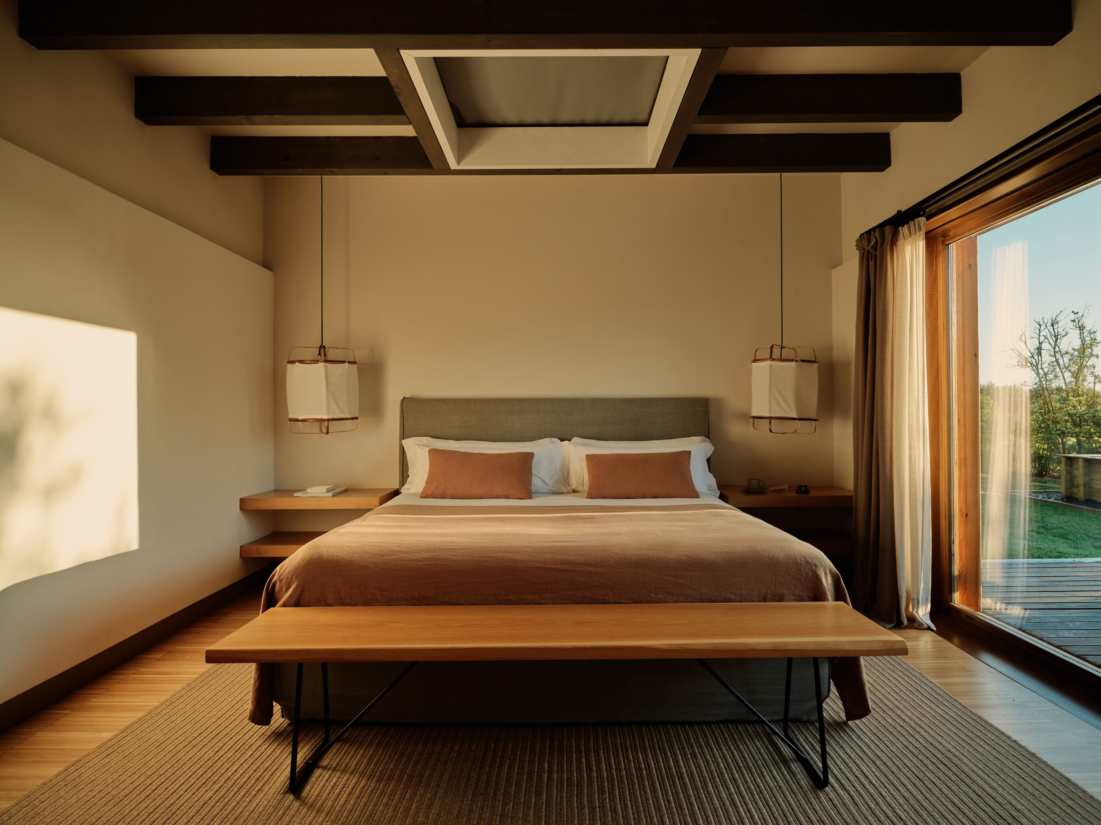 A modern bedroom with a large bed, bedside tables, a bench, and a view of the outdoors.