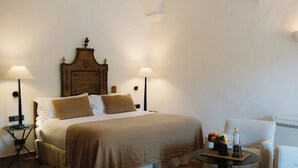 Een netjes opgemaakt bed met een houten hoofdbord, twee bedlampjes, een klein rond tafeltje met een fles en fruit, en een witte fauteuil.