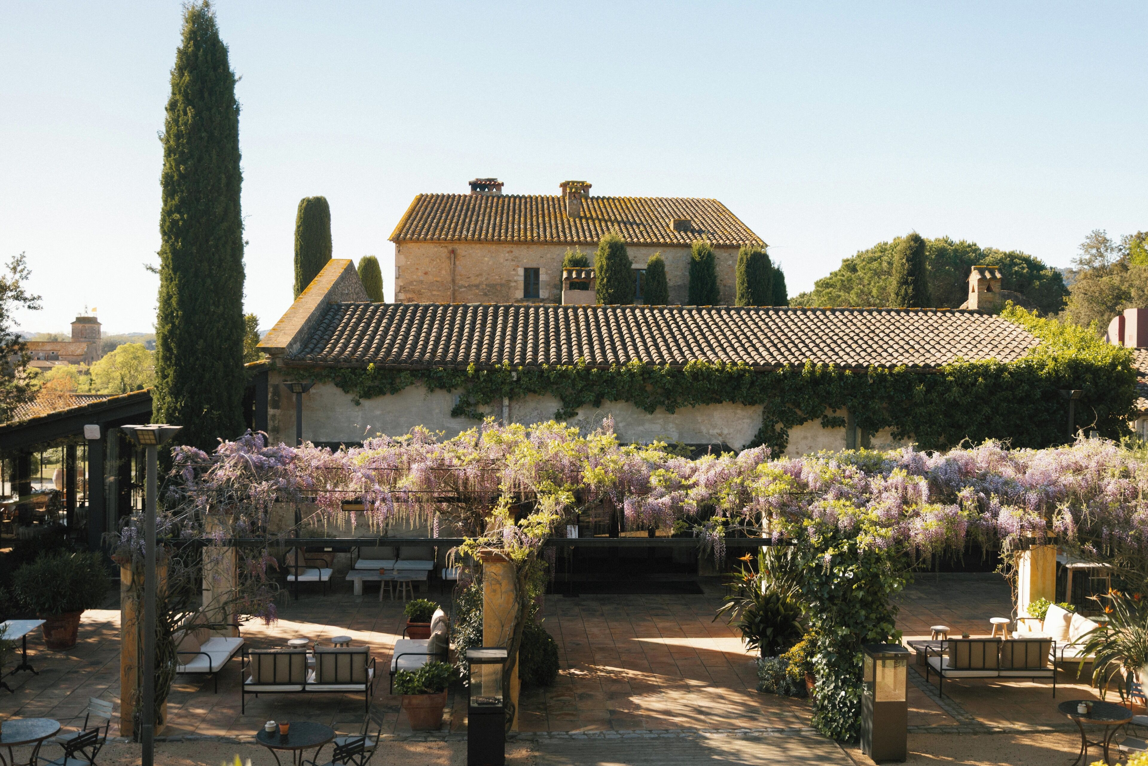 Une cour intérieure avec un bâtiment doté d’un toit en tuiles et d’une pergola recouverte de plantes grimpantes en fleurs.