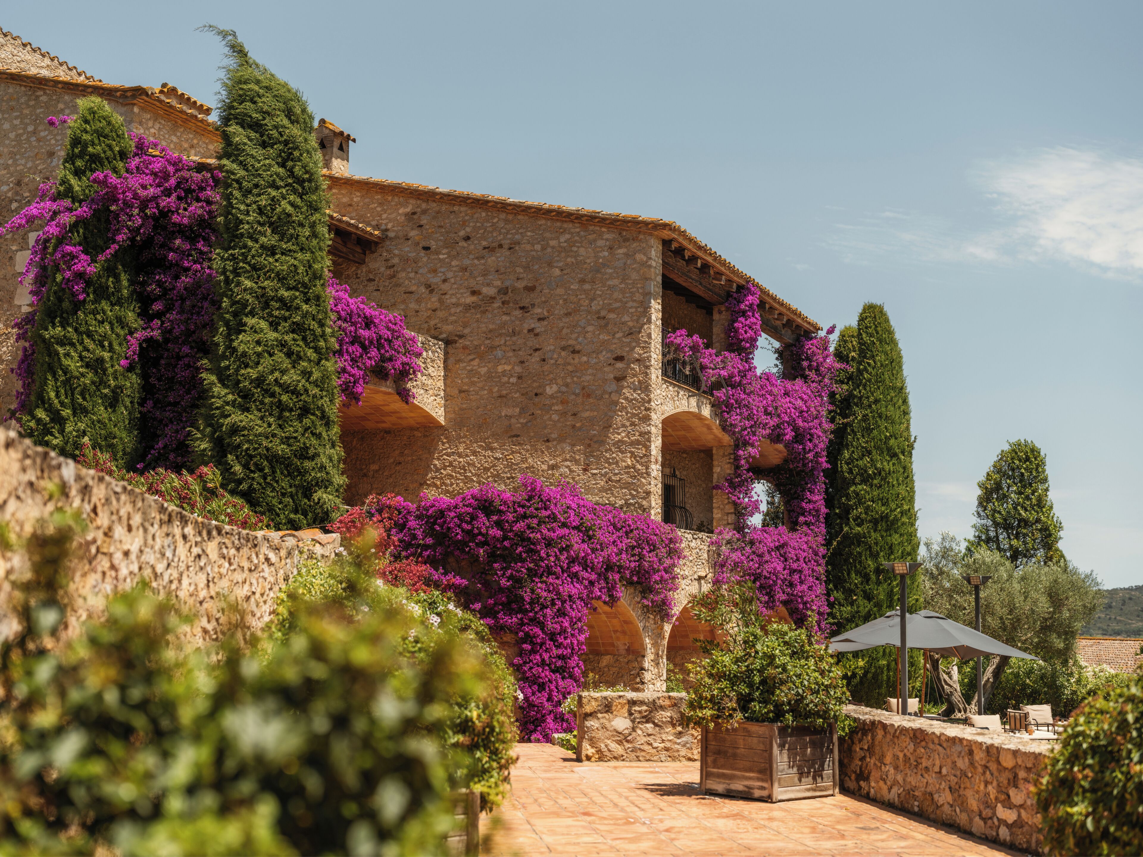 Un edificio de piedra con balcones cubiertos de flores moradas, un patio empedrado con mobiliario exterior y un cielo despejado.