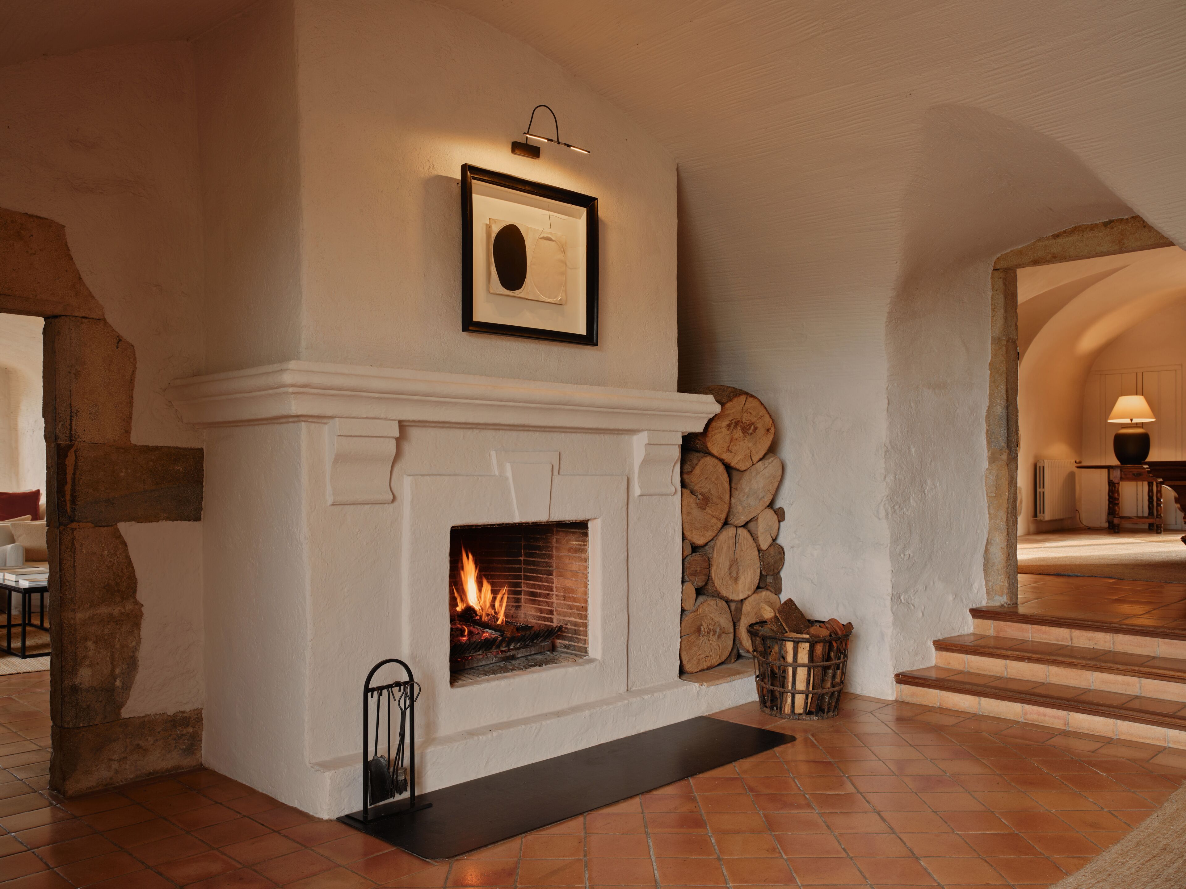 A fireplace with a fire, a framed picture above it, and a stack of logs to the right.