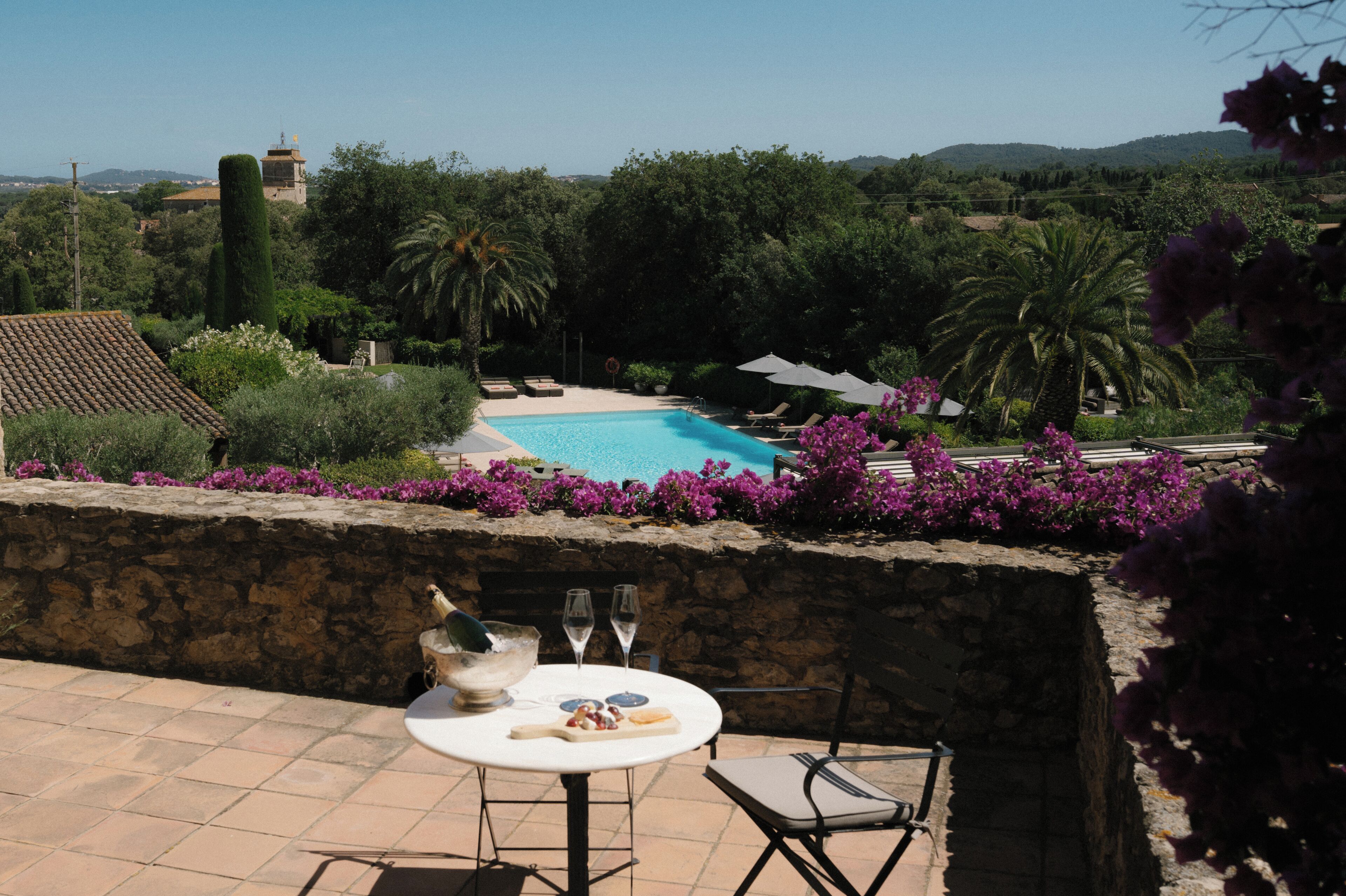 Zona junto a la piscina con un muro de piedra, una mesa con un cuenco y vasos, y vistas a la piscina y la vegetación circundante.
