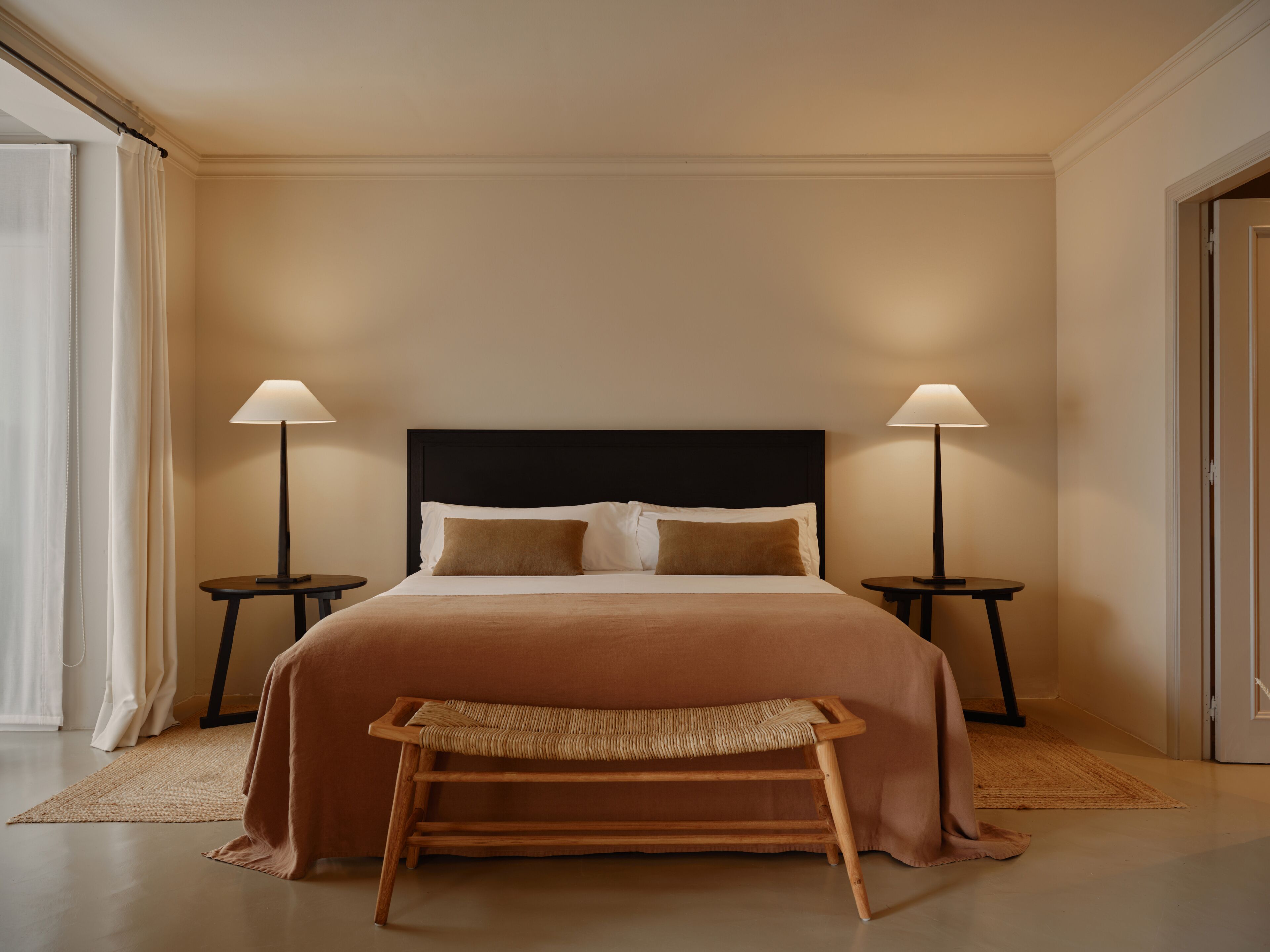 A hotel room with a large bed, two bedside tables, a bench, and a door.
