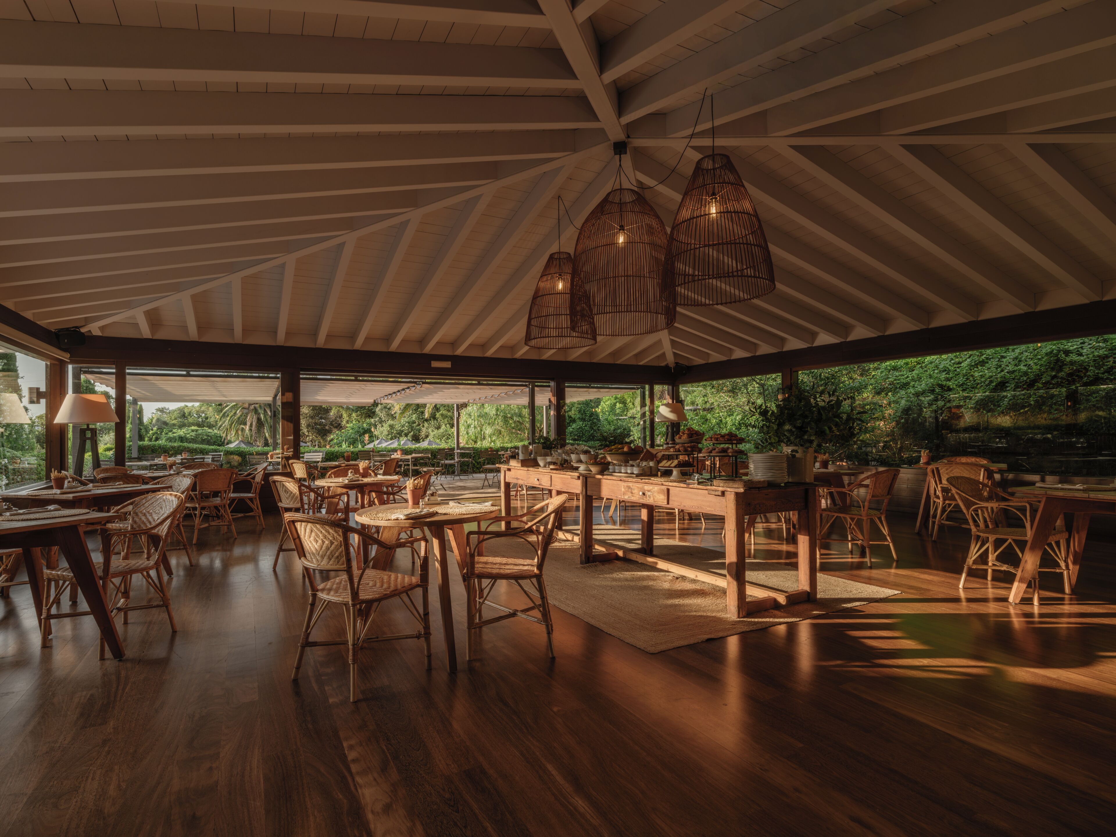 An open-plan dining area with wooden tables and chairs, large windows, and hanging light fixtures.