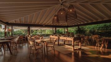 An open-plan dining area with wooden tables and chairs, large windows, and hanging light fixtures.