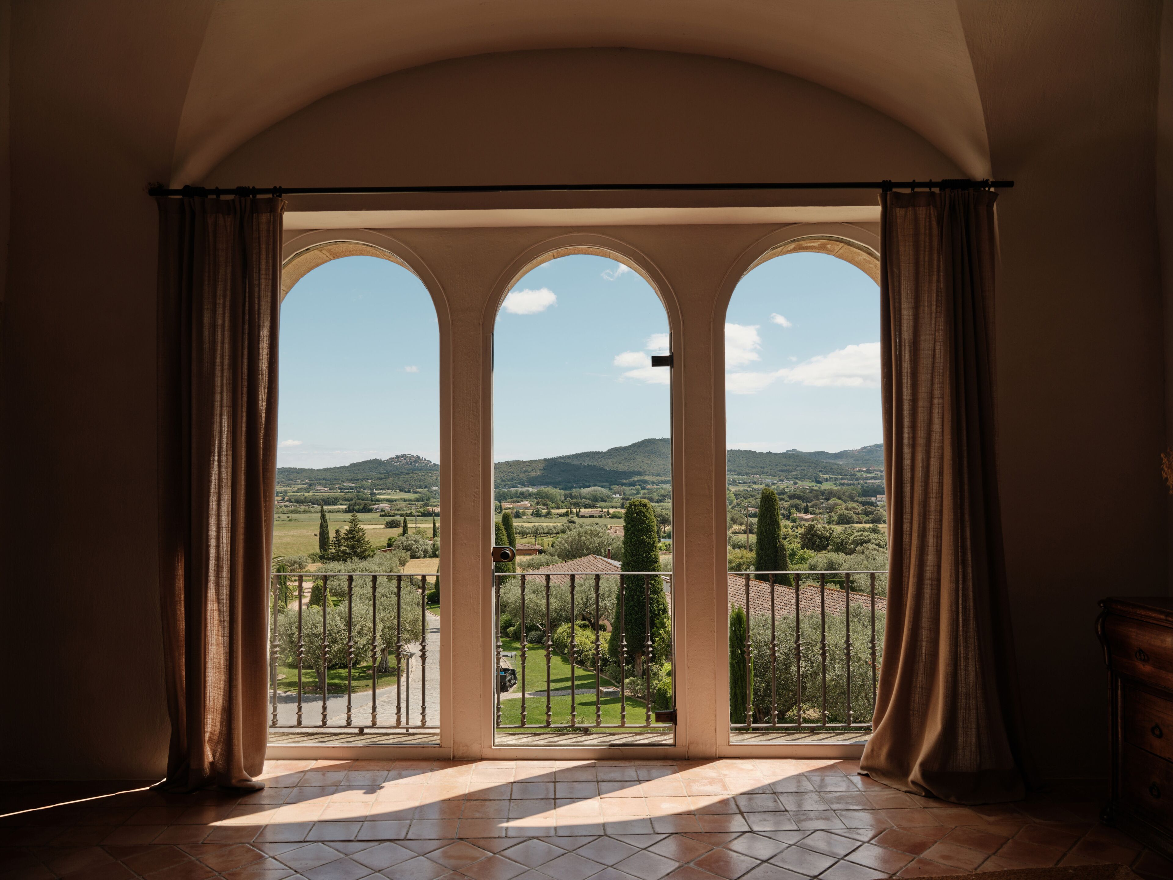 Una habitación con vistas a través de una ventana de tres arcos, un balcón con barandilla de metal y piso de baldosas.